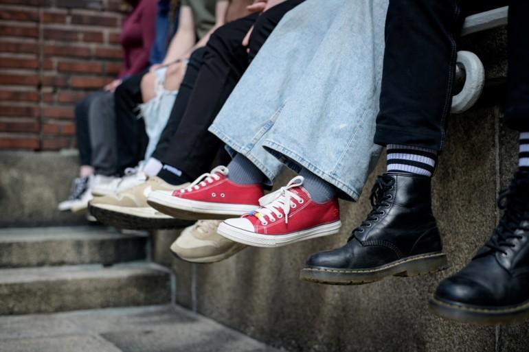 Kuvassa näkyy rivissä reunuksen päällä istuvien nuorten jalat ja erilaisia kenkiä.